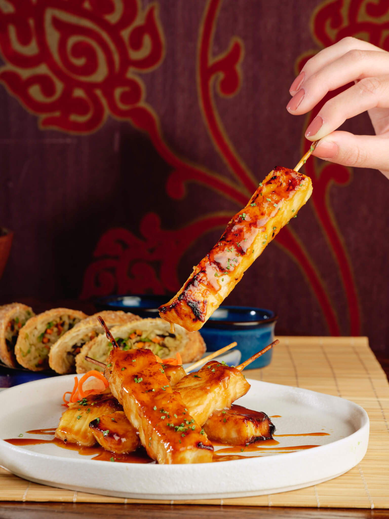person picking up a seabass satay skewers and egg rolls on plate in background