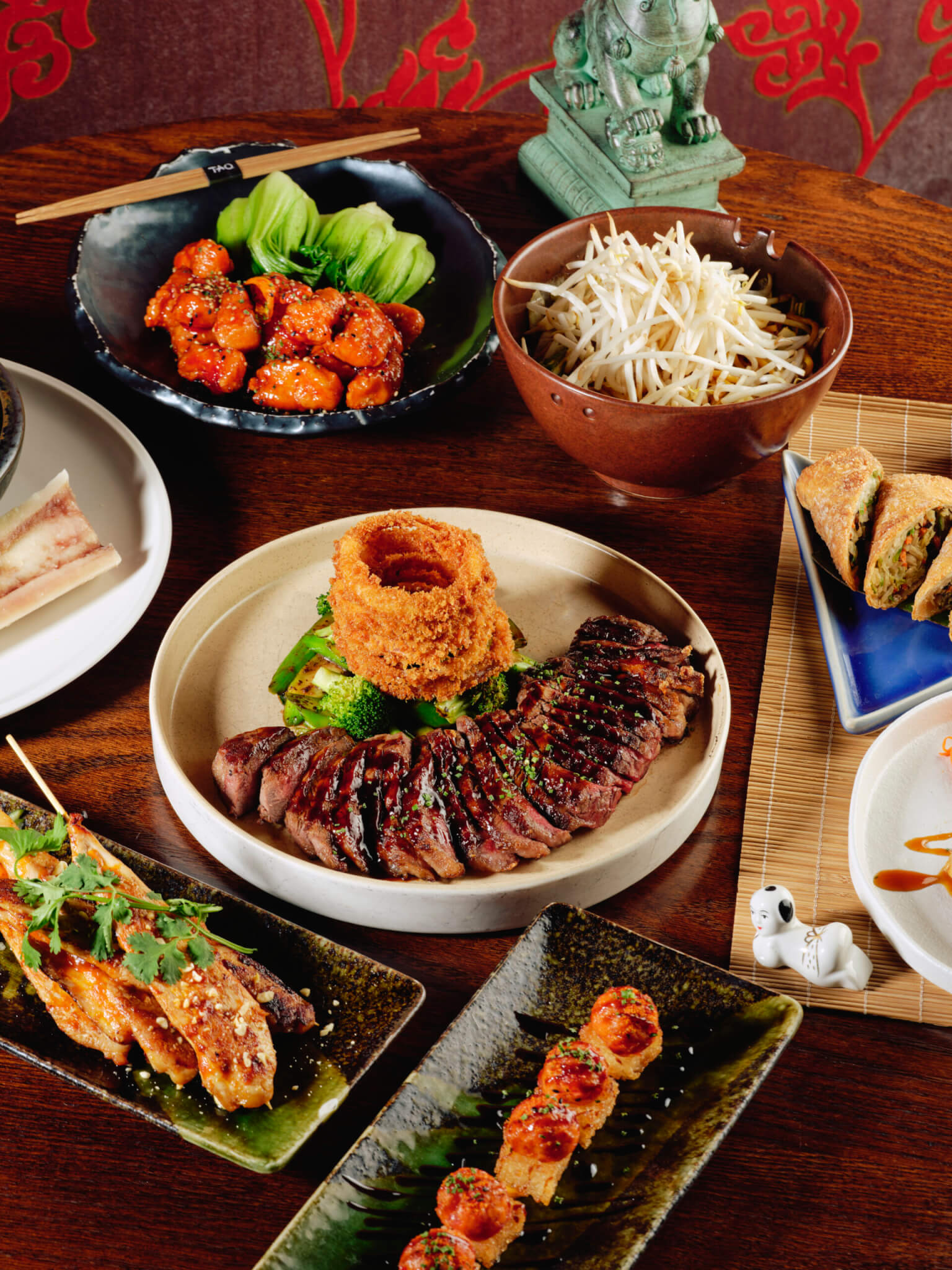 plat of tuna over crispy rice, plate of cut steak, brocolli and onion rings, bowl of bean sprouts, bowl of orange chicken with bok choy, plat of chicken satay skewers and bone marrow