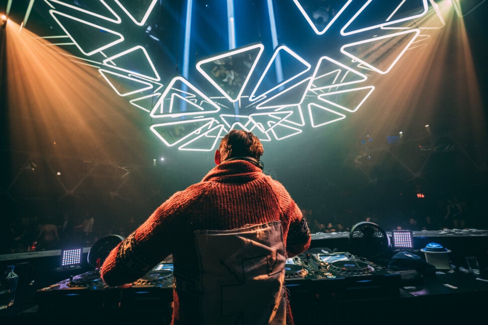 A DJ wearing a red sweater performs on stage in a nightclub, with geometric neon lights forming patterns overhead and a crowd visible in the dim background, setting the perfect scene for an unforgettable Party Favor experience.