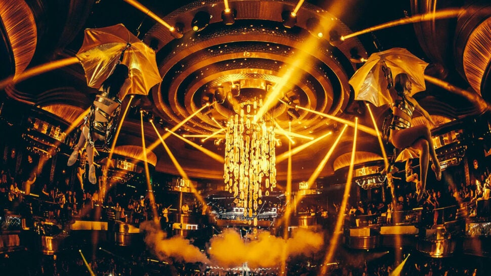 A nightclub filled with golden lights, lasers, smoke, and flying performers on stilts. The central chandelier shines as the crowd dances below in a vibrant, energetic atmosphere.