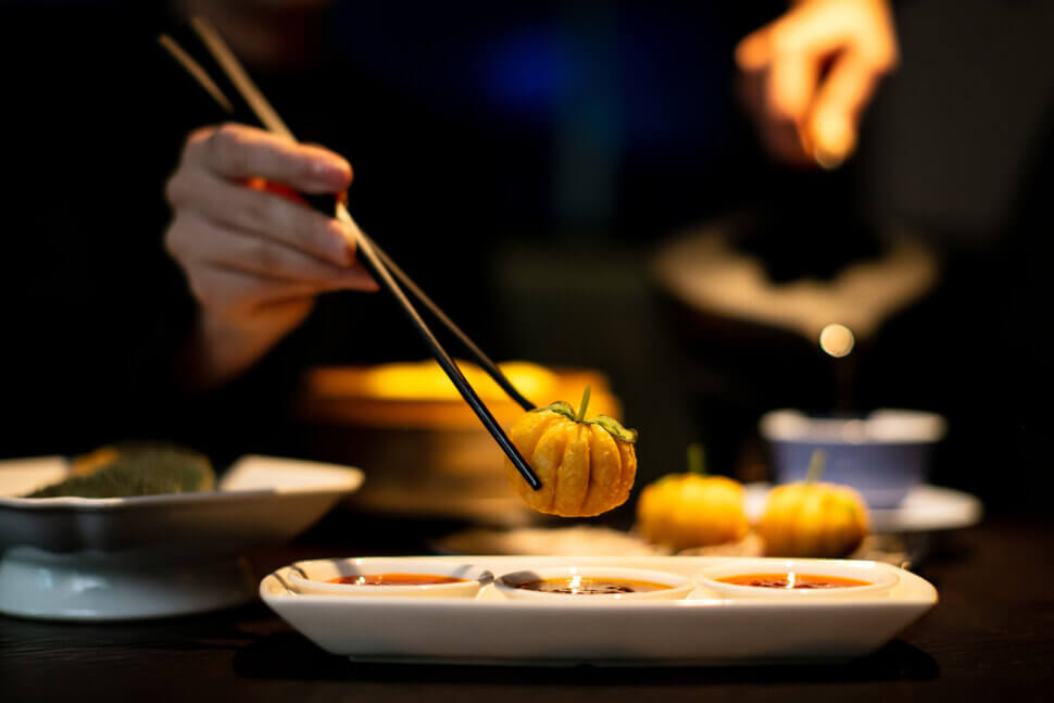 A person uses chopsticks to dip a small, pumpkin-shaped dim sum into sauce at Hakkasan Riyadh, with more food and sauces visible on the table in a dimly lit setting.