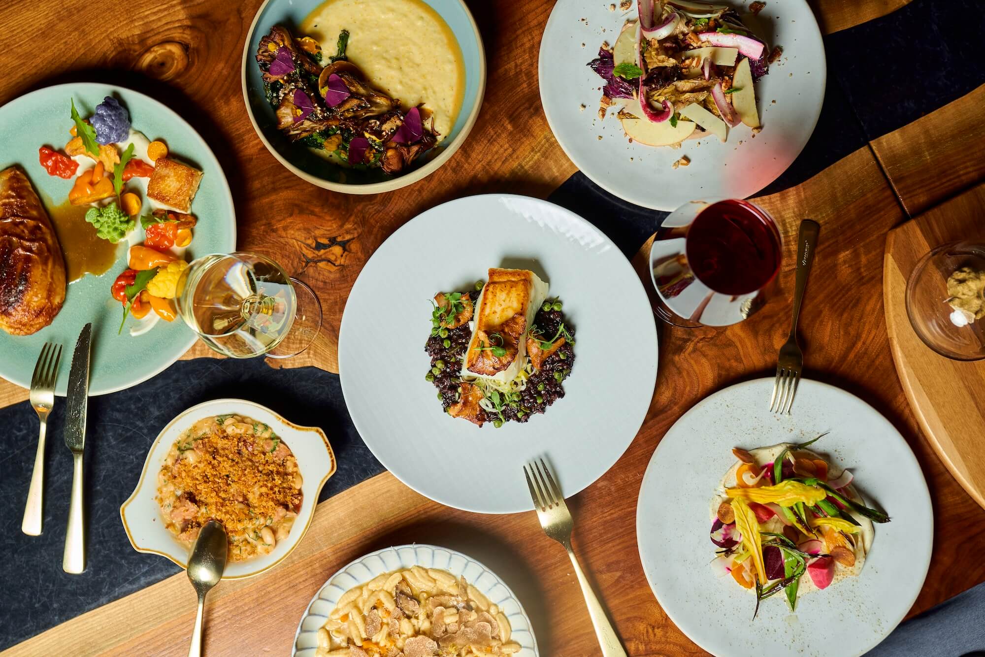A variety of gourmet dishes arranged on a wooden table. Plates include fish with greens, pasta, colorful vegetables, and a dessert. There are also glasses of red and white wine, showcasing an elegant dining setting.
