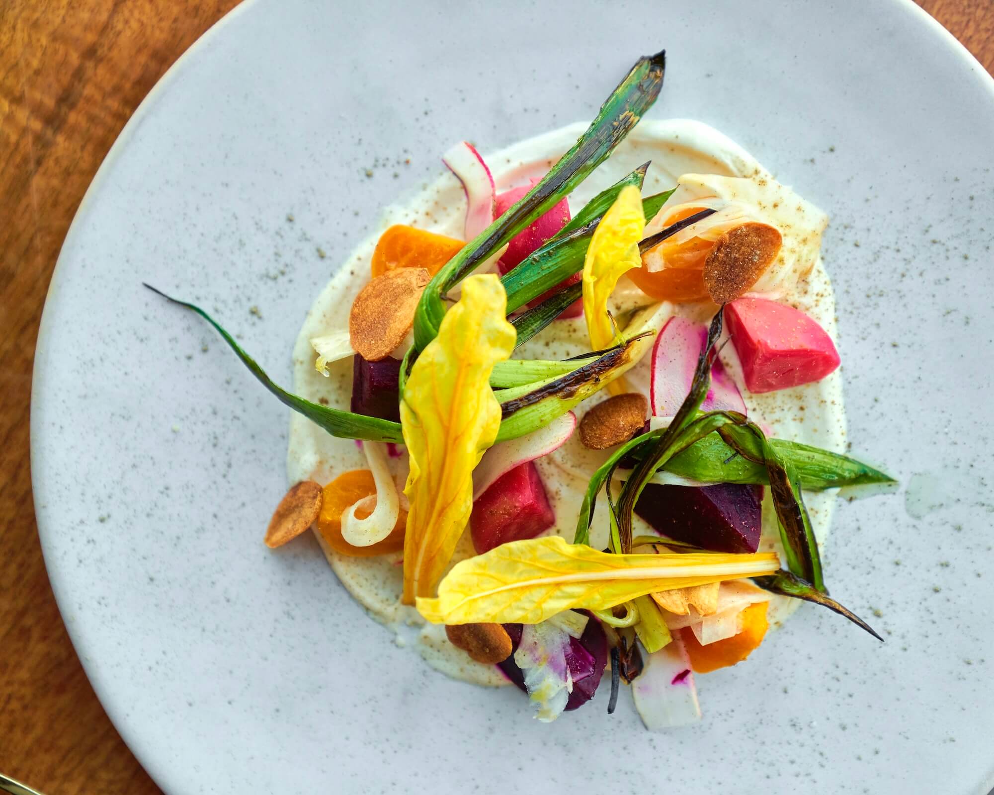 A colorful dish elegantly arranged on a white plate, featuring a variety of fresh vegetables like beets, spring onions, and radishes, garnished with vibrant yellow flower petals and thinly sliced pieces, served on a creamy white sauce.