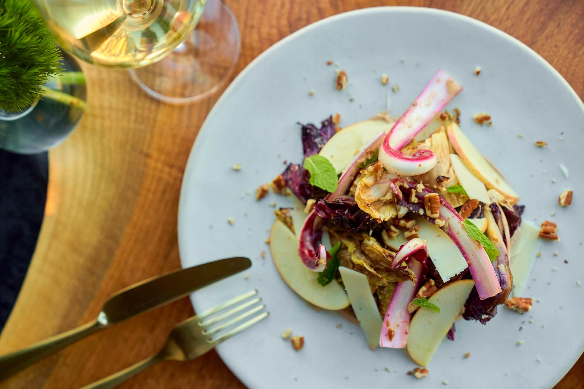 A gourmet salad with endive, apple slices, nuts, and cheese on a white plate. A fork and knife are placed beside the plate on a wooden table, with a glass of white wine nearby.