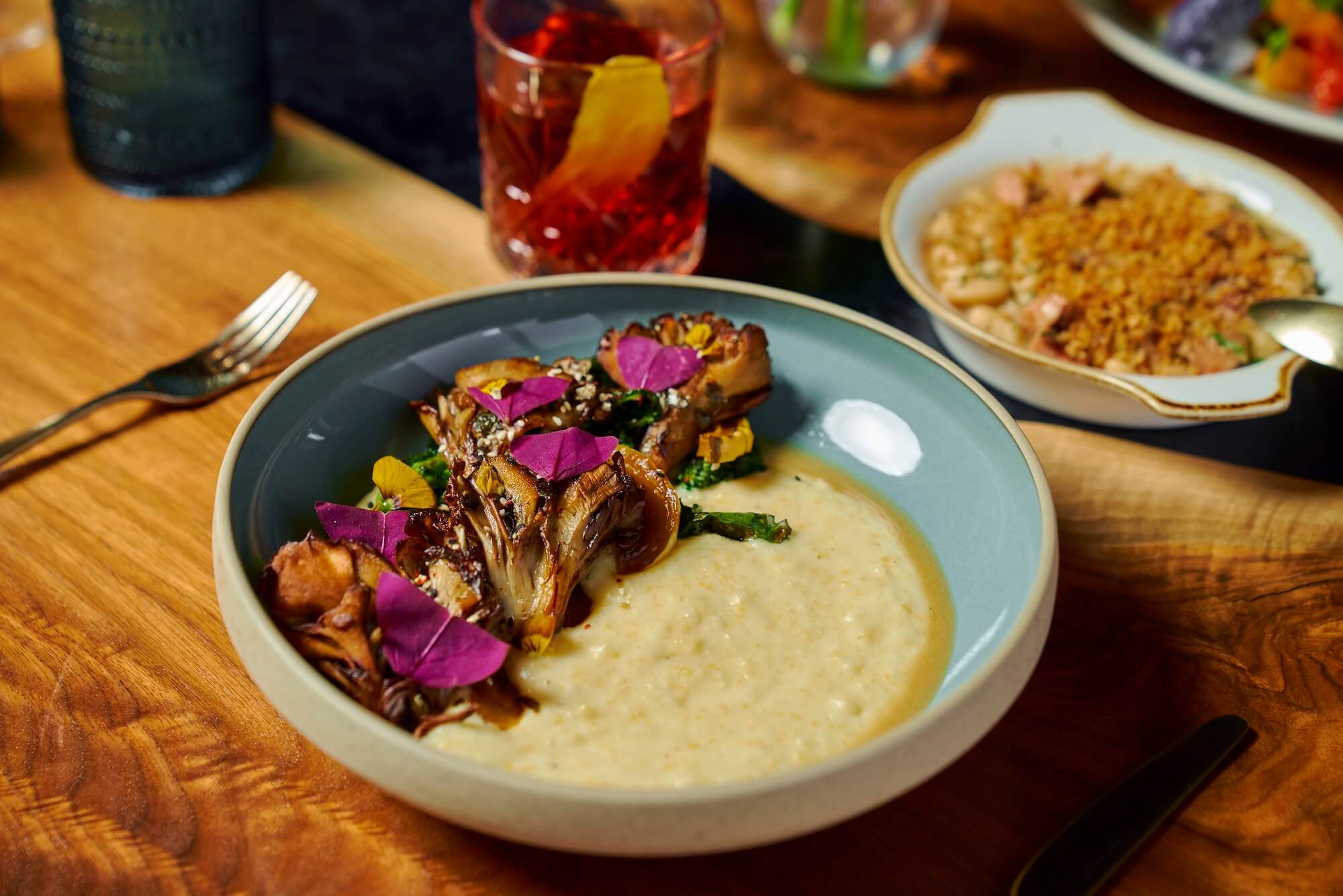 A bowl of creamy polenta topped with roasted mushrooms and purple edible flowers. A reddish drink with an orange slice is in the background, along with a dish of beans and grains. The setting is on a wooden table with cutlery.