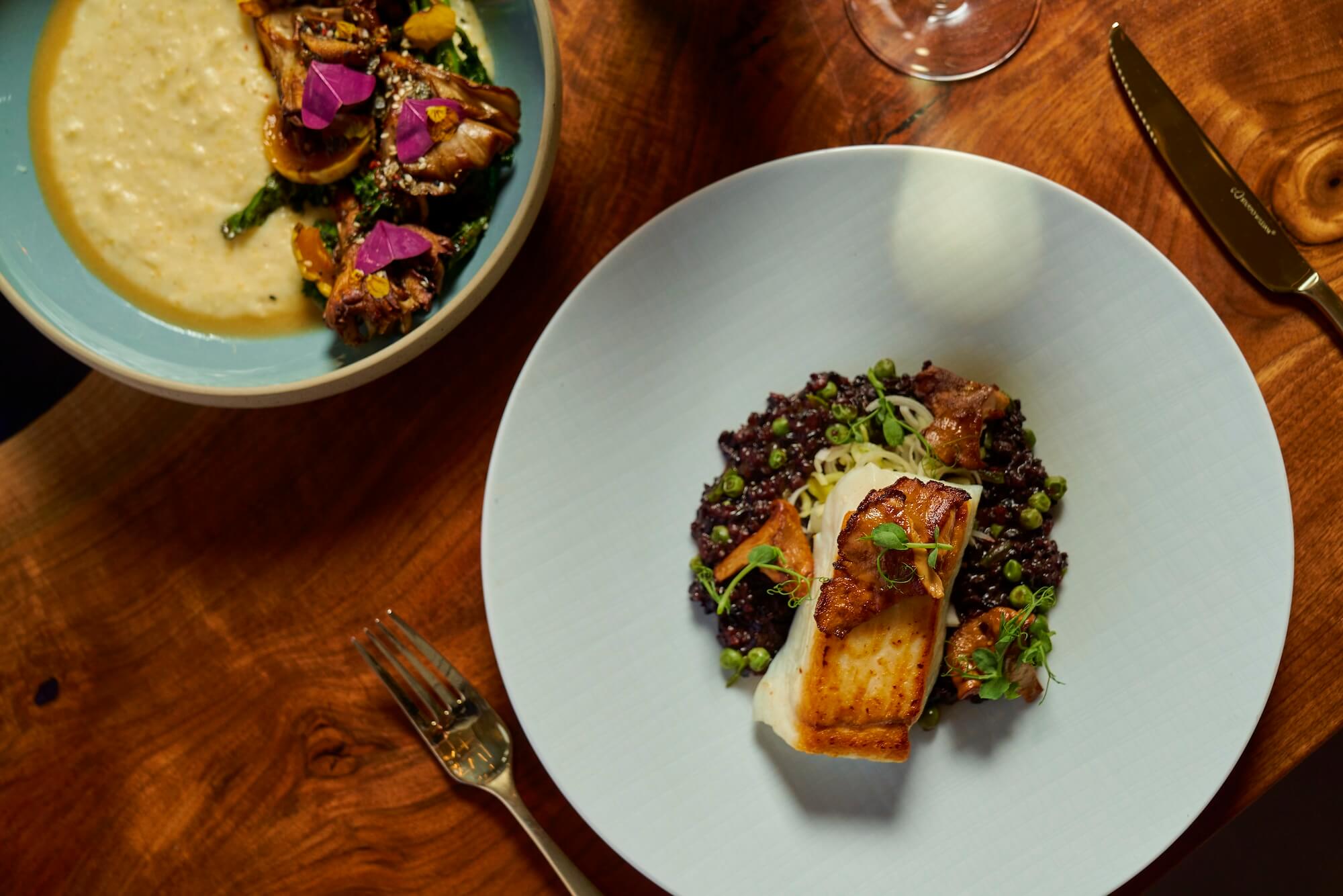 A wooden table with two plates of gourmet food: one with a dish featuring seared fish on grains with herbs, and another with a creamy dish topped with artichokes and colorful garnishes. Nearby are a fork and a wine glass.