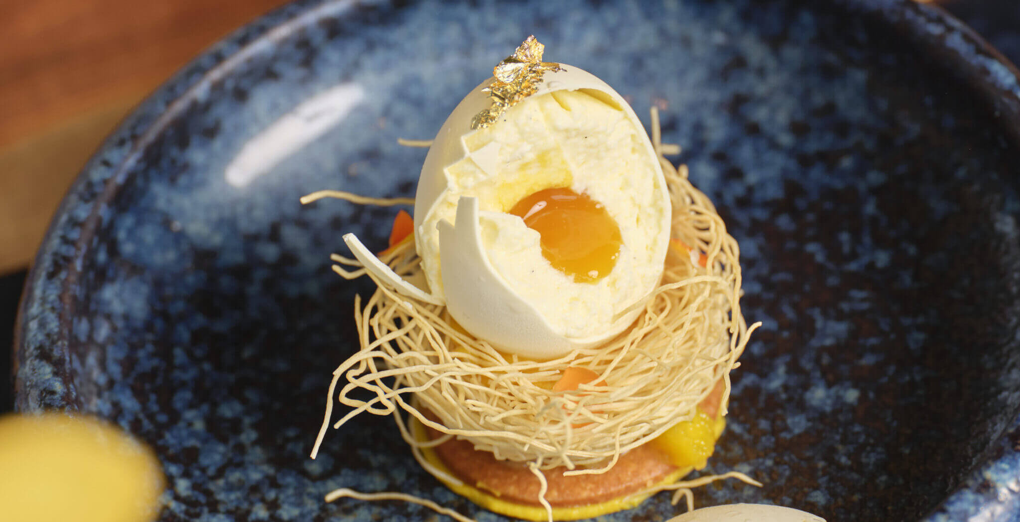 A gourmet dessert resembling a poached egg sits atop a nest of spun sugar on a blue ceramic plate. The dessert has an outer shell, creamy filling, and a yolk-like center, accented with a small piece of gold leaf.
