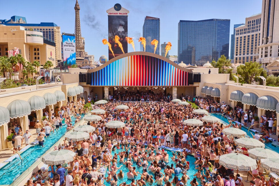 A large crowd enjoys a lively pool party with umbrellas, surrounded by tall buildings and fire effects, near Caesars Palace in Las Vegas on a sunny day—capturing the vibrant energy of Marquee Dayclub Private Events.