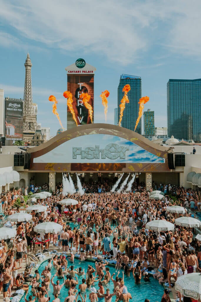 A large crowd at a Las Vegas pool party with people dancing, water spray, fire jets, and city buildings in the background—including replicas of famous landmarks—captures the vibrant atmosphere of Marquee Dayclub Private Events.
